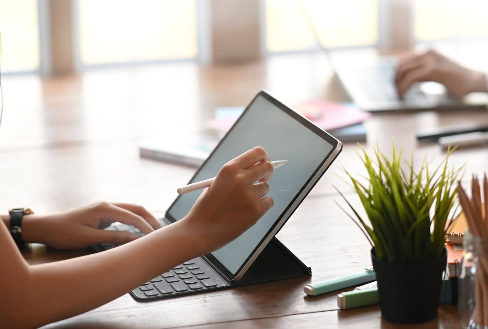 someone using a touchscreen device with a pen. plants and stationary are scattered on the table